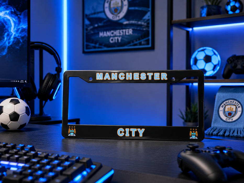 A black license plate frame featuring 'MANCHESTER CITY' in white and gold lettering, displayed on a gaming desk with blue LED lighting, a keyboard, controller, soccer ball, and Manchester City merchandise.