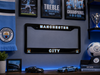 A black Manchester City license plate frame mounted on a blue wall, surrounded by framed Manchester City football memorabilia, a scarf, trophies, a soccer ball, and small car models.