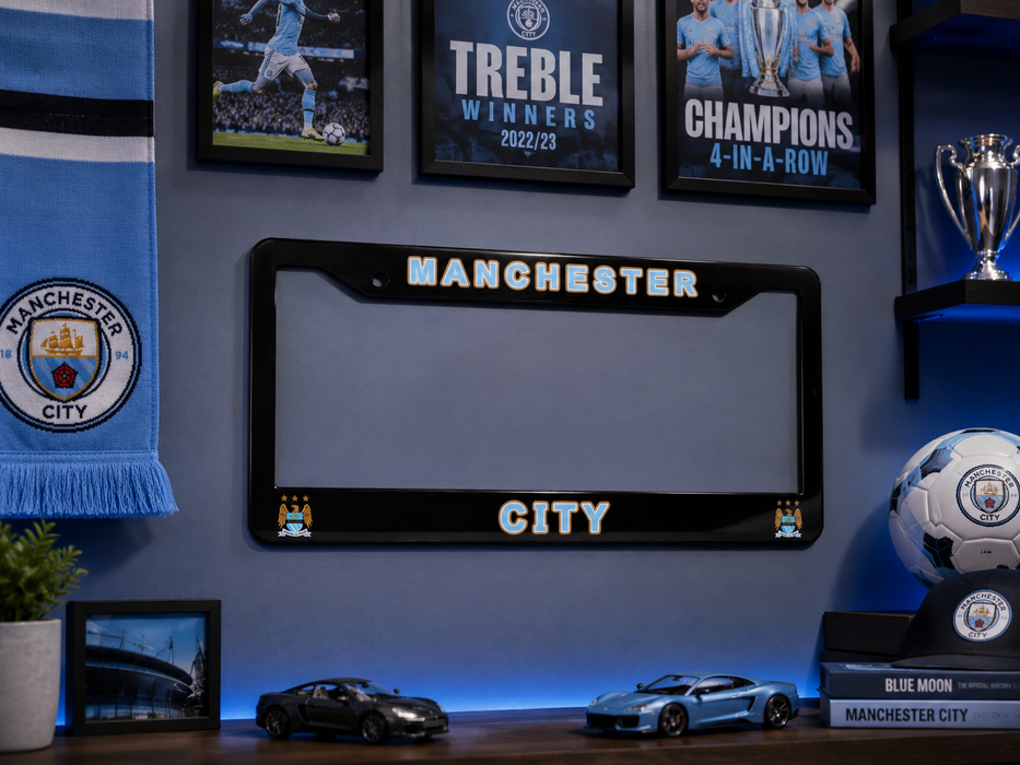 A black Manchester City license plate frame mounted on a blue wall, surrounded by framed Manchester City football memorabilia, a scarf, trophies, a soccer ball, and small car models.