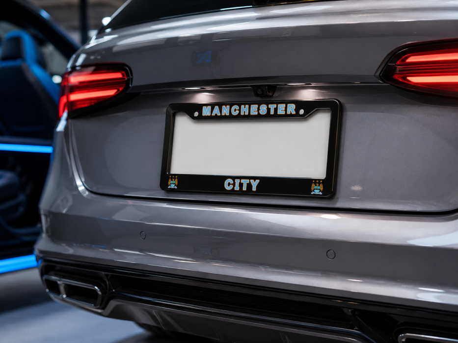 Close-up rear view of a gray car showing a black license plate frame with the words “MANCHESTER” at the top and “CITY” at the bottom, mounted on the license plate area.