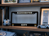 A black Manchester City license plate frame with gold and white lettering, displayed on a wooden shelf alongside a soccer ball, framed ticket, and a toy car.