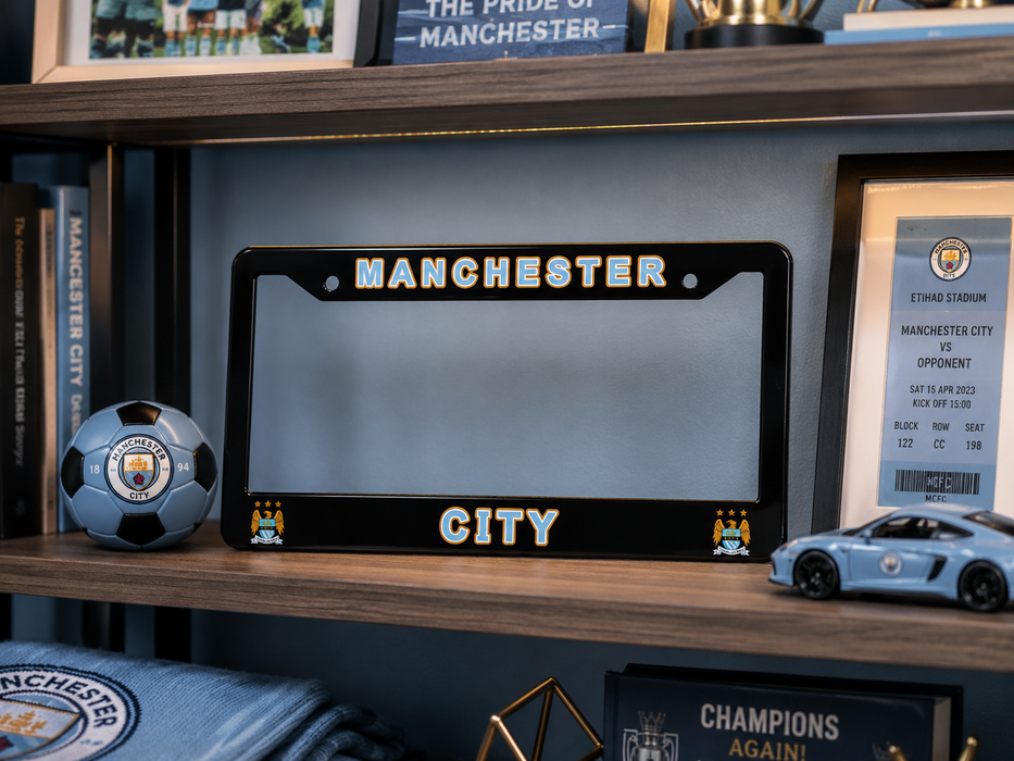 A black Manchester City license plate frame with gold and white lettering, displayed on a wooden shelf alongside a soccer ball, framed ticket, and a toy car.