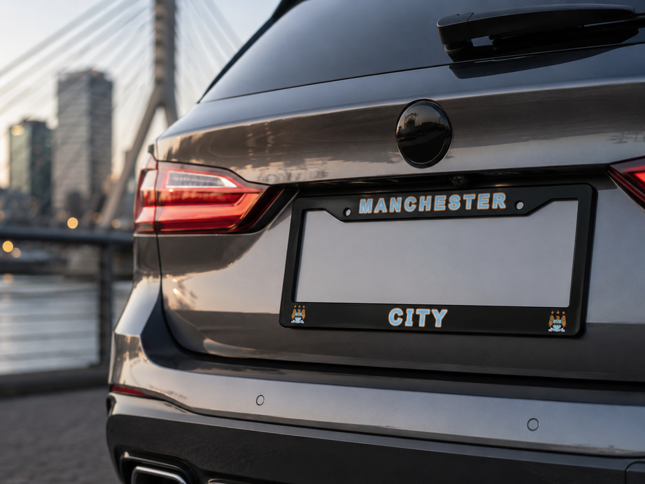 Close-up rear view of a dark gray SUV showing a custom license plate frame with 'MANCHESTER' at the top and 'CITY' at the bottom, set against a blurred cityscape background.
