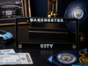 A black Manchester City license plate frame displayed on a dark tabletop, surrounded by soccer-themed merchandise including a blue Manchester City soccer ball, club tickets, a crest patch, a small car model, and framed stadium memorabilia.
