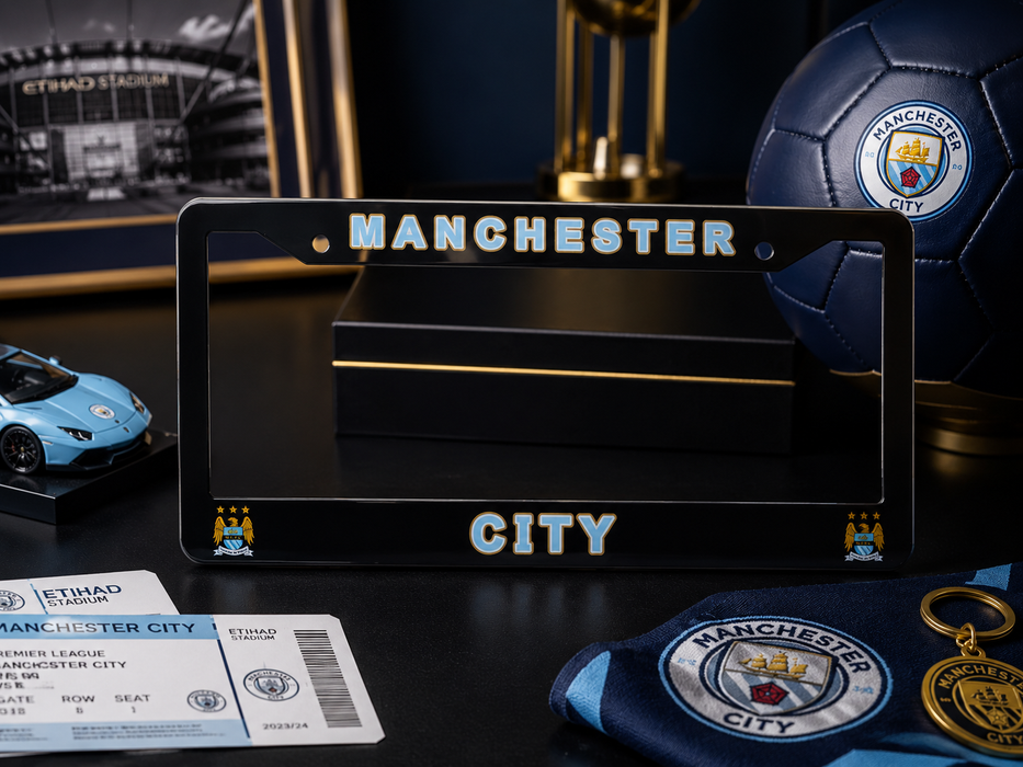 A black Manchester City license plate frame displayed on a dark tabletop, surrounded by soccer-themed merchandise including a blue Manchester City soccer ball, club tickets, a crest patch, a small car model, and framed stadium memorabilia.