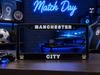 A black license plate frame featuring Manchester City branding, including 'MANCHESTER' at the top, 'CITY' at the bottom, and team crest details in the corners, displayed on a wooden desk in a gaming room setting.