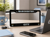 A black license plate frame with Manchester City branding, displayed on a wooden desk with a laptop, phone, mug, and other office items in a bright room.