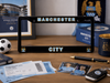 A black Manchester City license plate frame displayed on a wooden desk with football-themed items, including a ticket, cards, a pen, and a key fob. The frame features 'MANCHESTER' at the top and 'CITY' at the bottom in light blue with a gold outline, flanked by the Manchester City crest.