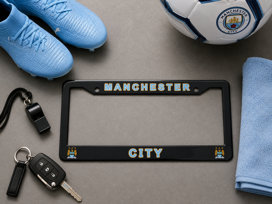 A black Manchester City license plate frame with gold and light blue lettering, styled in a flat lay with a Manchester City soccer ball, light blue cleats, keys, a whistle, and a towel on a gray surface.