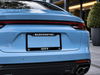 A light blue Porsche rear view with a black license plate frame featuring the words 'MANCHESTER CITY' in white. The car is parked outdoors in front of a modern building.