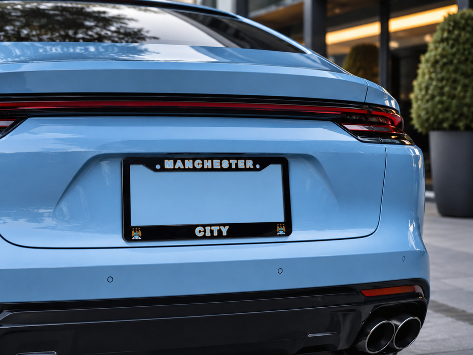 A light blue Porsche rear view with a black license plate frame featuring the words 'MANCHESTER CITY' in white. The car is parked outdoors in front of a modern building.