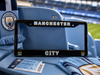 A black Manchester City license plate frame with gold and white lettering, displayed on a light blue stadium seat alongside a Manchester City scarf and ticket.