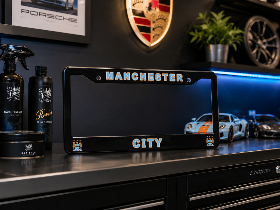 A black license plate frame with 'MANCHESTER CITY' in silver lettering, displayed on a dark workbench in a garage setting with a Porsche crest on the wall behind it.