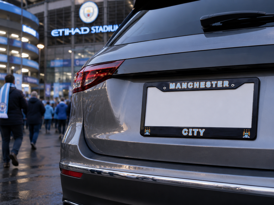 A close-up rear view of a silver car with a black license plate frame featuring the words 'MANCHESTER CITY' and small team crests in the corners. The background shows a blurred Etihad Stadium entrance with people walking outside.