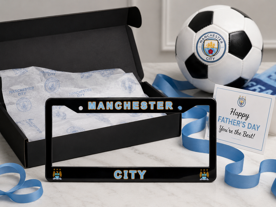 A Manchester City-themed Father's Day gift set featuring a black license plate frame with gold and blue lettering, a black gift box with Manchester City tissue paper, a soccer ball, a 'Happy Father's Day' card, and blue ribbons on a white marble surface.