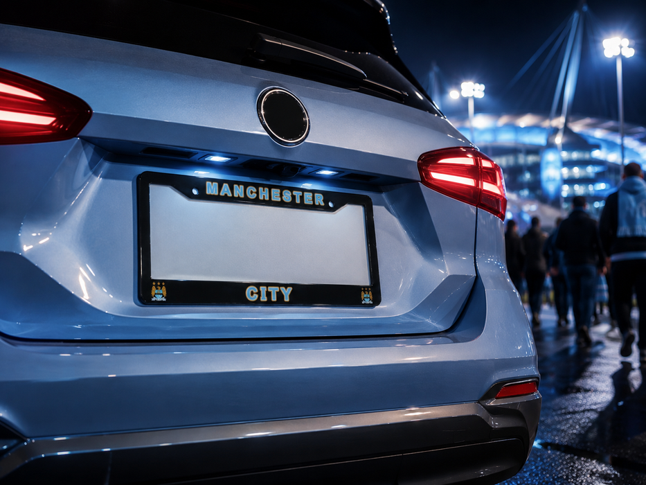 A close-up rear view of a silver car at night with a black license plate frame featuring the words 'MANCHESTER' at the top and 'CITY' at the bottom.