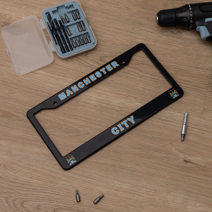 Black license plate frame with 'Manchester City' text on a wooden surface, surrounded by tools.