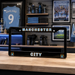 Manchester City-themed license plate on a wooden surface with sports memorabilia in the background.