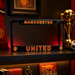 Manchester United license plate frame with club logo and text on a shelf with sports memorabilia.