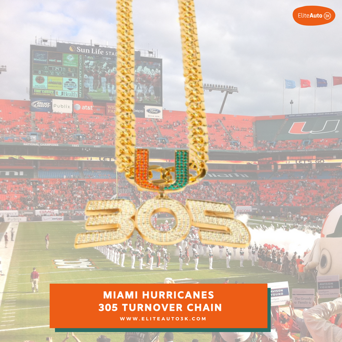 A gold Miami Hurricanes 305 turnover chain necklace with a 'U' pendant embellished in orange and green, and '305' in rhinestones, shown against a blurred background of a football stadium.