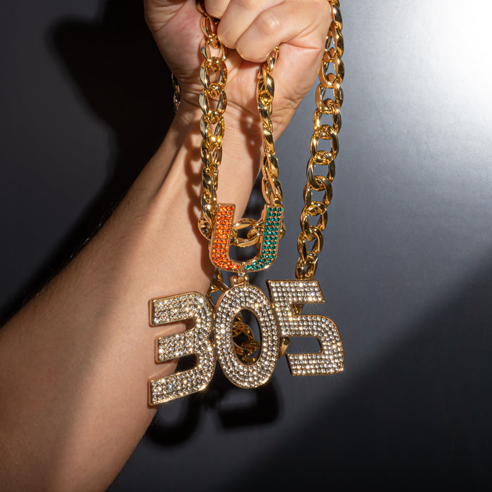 A close-up of a hand holding a chunky gold chain necklace with a large '305' pendant covered in clear rhinestones, alongside two smaller letter pendants in orange and green.