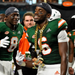 Two Miami Hurricanes football players in green jerseys celebrate on the field, with one wearing a large gold chain and a small alligator charm. Visible Adidas logos appear on the uniforms, and an ElnaAuto badge is in the top-right corner.