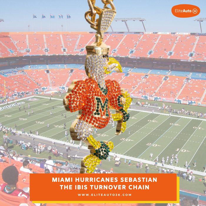 A close-up of a gold-toned chain with a Miami Hurricanes football player charm, shown in front of a blurred stadium background. The charm features a gold eagle head and a football-shaped body covered in orange, green, and white rhinestones, with the letter 'M' on the front.