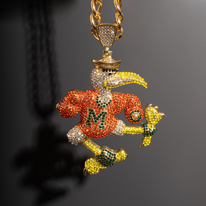 A gold-toned pendant necklace featuring a highly detailed, rhinestone-encrusted mascot character in orange, yellow, green, and white, suspended from a thick gold chain against a dark gray background.
