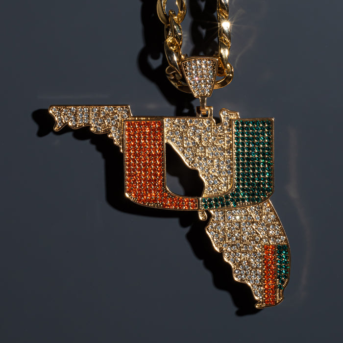 Gold-tone pendant necklace shaped like the state of Florida, heavily embellished with clear rhinestones and featuring orange, green, and gold sections, shown on a dark gray background.