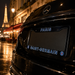 A close-up rear view of a black Mercedes-Benz car on a wet city street at night, with Paris-themed license plate decals reading "PARIS" and "SAINT-GERMAIN" and a PSG logo, against a blurred background of the Eiffel Tower and lit buildings.