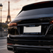 A close-up rear view of a glossy black SUV parked outdoors in Paris, with the Eiffel Tower blurred in the background. The vehicle has a custom license plate frame holding a white plate with blue text reading 'PARIS' at the top and 'SAINT-GERMAIN' at the bottom.