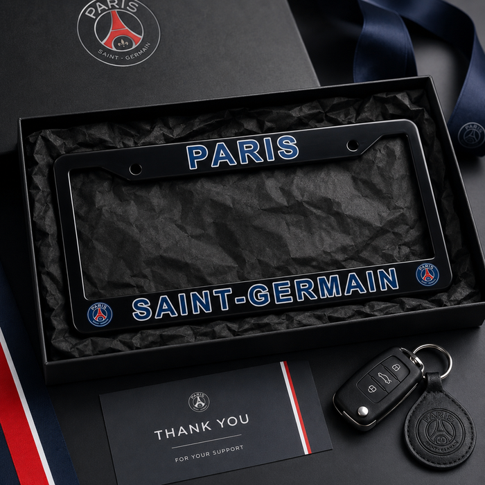 A black Paris Saint-Germain license plate frame with blue and white lettering, presented in a black gift box with a crumpled black paper insert. A car key fob and a thank-you card are placed beside it on a dark surface, with a Paris Saint-Germain crest visible on the box lid and key fob.