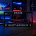 A black Paris Saint-Germain license plate frame on a desk in a gaming room setup, with PSG logos and 'PARIS SAINT-GERMAIN' text, alongside a keyboard, monitor, and game controller.