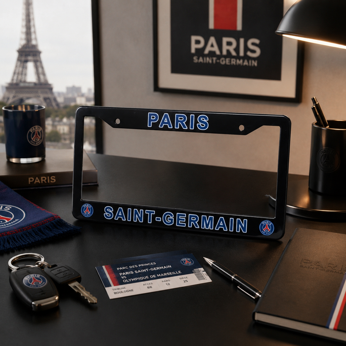 A Paris Saint-Germain license plate frame displayed on a dark desk, surrounded by Paris-themed items including a mug, notebook, pen, and a card. The frame features 'PARIS' at the top and 'SAINT-GERMAIN' at the bottom in blue lettering with PSG logos on the sides.