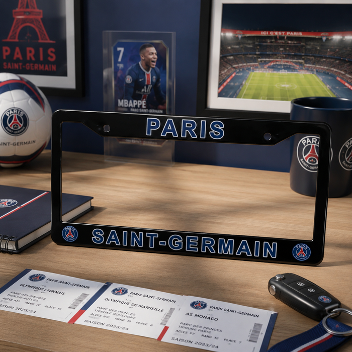 A Paris Saint-Germain license plate frame displayed on a wooden desk, surrounded by Paris football-themed items including a PSG soccer ball, tickets, a mug, and framed prints.