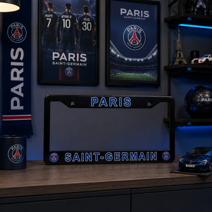 A Paris Saint-Germain license plate frame displayed on a wooden desk in a themed room. The black frame features 'PARIS' in blue at the top and 'SAINT-GERMAIN' in blue at the bottom, flanked by the club's crest.