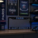 A Paris Saint-Germain license plate frame displayed on a wooden desk in a themed room. The black frame features 'PARIS' in blue at the top and 'SAINT-GERMAIN' in blue at the bottom, flanked by the club's crest.
