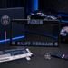 A Paris Saint-Germain license plate frame set displayed on a dark surface, including the black license plate frame with blue and white 'PARIS SAINT-GERMAIN' text, a branded Paris Saint-Germain box, a ticket, a keychain, a ribbon, and a soccer ball. A blue sports car is visible in the background.