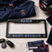 A Paris Saint-Germain license plate frame in black with blue and red lettering, styled in a flat lay with a navy jacket, soccer ball, keys, scarf, and tickets on a white marble background.