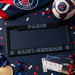 A black Paris Saint-Germain license plate frame with blue and white lettering, styled on a dark blue background with a Paris FC scarf, soccer ball, tickets, confetti, and a gold medal.