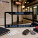 A black license plate frame with 'PARIS' and 'SAINT-GERMAIN' in blue lettering, displayed on a table with Paris-themed travel props including a passport, ticket, car key fob, and a Paris Saint-Germain keychain. The background shows a blurred Paris Metro station interior.