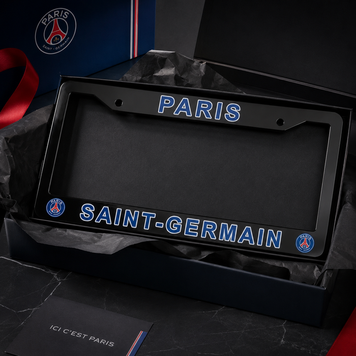 A black Paris Saint-Germain license plate frame displayed inside its branded presentation box, with 'PARIS' at the top and 'SAINT-GERMAIN' at the bottom in blue outlined lettering, plus PSG team logos in the corners.