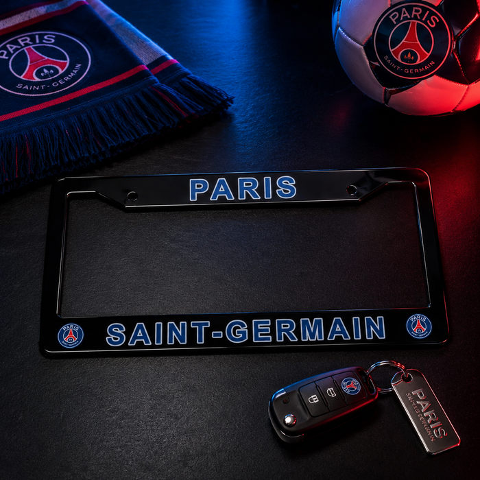 A black Paris Saint-Germain license plate frame with blue and red lettering, displayed on a dark surface with a car key fob, a Paris soccer ball, and a PSG scarf in the background.