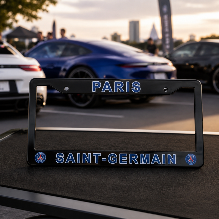 A black license plate frame with 'PARIS' on the top and 'SAINT-GERMAIN' on the bottom, featuring blue lettering and PSG club logos, displayed outdoors on a table with blurred cars in the background.