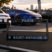 A black license plate frame with 'PARIS' on the top and 'SAINT-GERMAIN' on the bottom, featuring blue lettering and PSG club logos, displayed outdoors on a table with blurred cars in the background.
