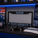 A Paris Saint-Germain license plate frame displayed on a dark shelf with blue LED lighting, alongside a small black sports car model, a Paris FC ticket, and team merchandise.
