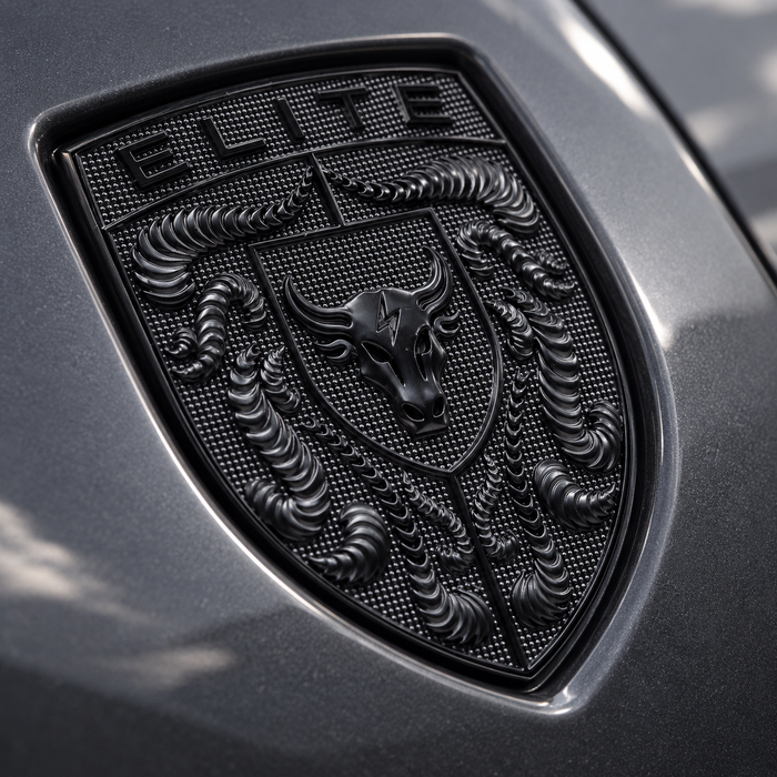 A close-up of a black porsche shield emblem on a dark metallic gray vehicle surface, featuring the word ELITE at the top and a bull head in the center.