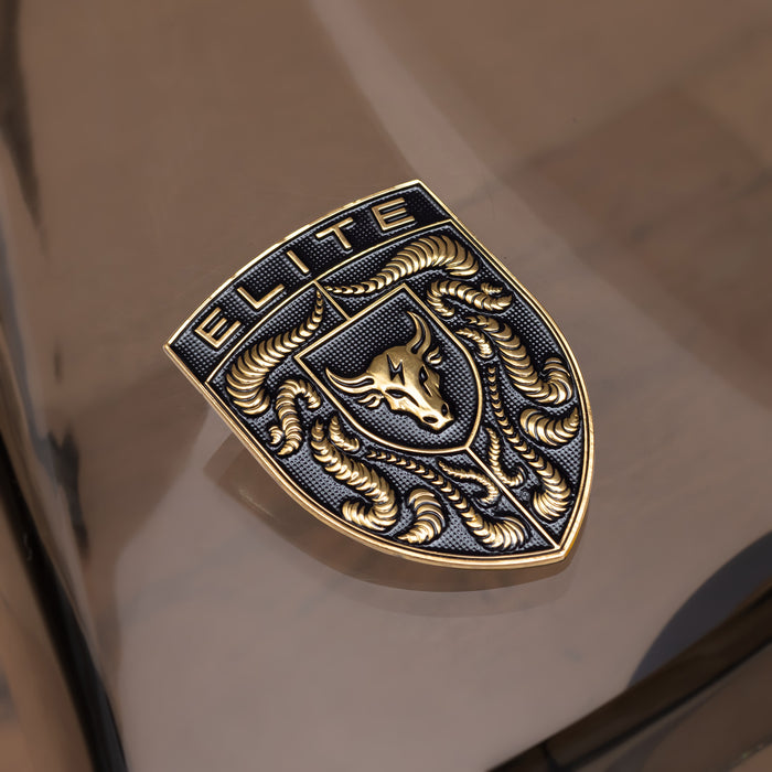 A close-up of a metallic porsche badge or emblem with the word "ELITE" at the top, featuring a stylized bull head and crumrine designs in polished gold-toned metal over a dark textured background.