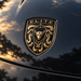 A close-up of a metallic gold and black porsche emblem featuring a bull head and the word 'ELITE' at the top, mounted on a glossy dark car body with reflections of trees and sky.