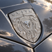 A close-up of a metallic Chrome Silver Brahma Bull Hood Emblem featuring a shield with a bull head and the word "ELITE" at the top, mounted on a dark gray vehicle hood with sunlight and tree reflections.